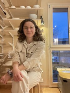 Lilly Haas sitting in her ceramic studio with all over her pieces on a shelf behind her as she smiles for the camera