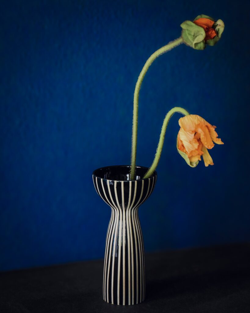 Keramik bemalen mit fortgeschrittenem Streifen-Design: Schlanke schwarz-weiße Vase mit feinen vertikalen Linien und zwei orangefarbenen Blüten vor dunklem Hintergrund.