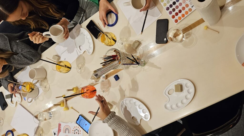 Keramik Malkurs: Teilnehmer:innen sitzen gemeinsam am Tisch und gestalten Tassen und Teller mit Pinseln, Glasuren und Klebeband, während Getränke daneben stehen.