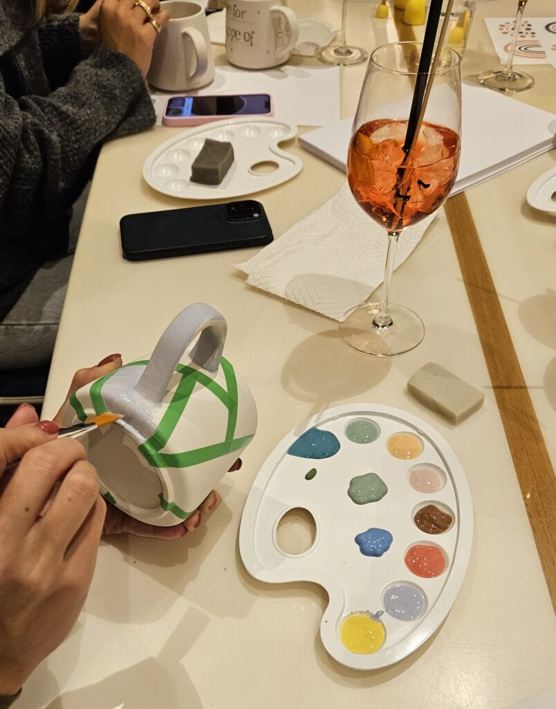 Keramik bemalen mit der Klebeband-Methode: Eine Tasse wird mit grünem Tape abgeklebt und mit Glasur aus einer Farbpalette bemalt, um klare geometrische Linien zu gestalten.