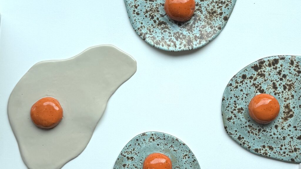 Handmade ceramic egg sculptures in speckled teal glaze and off white color with orange yolks, displayed on a white surface