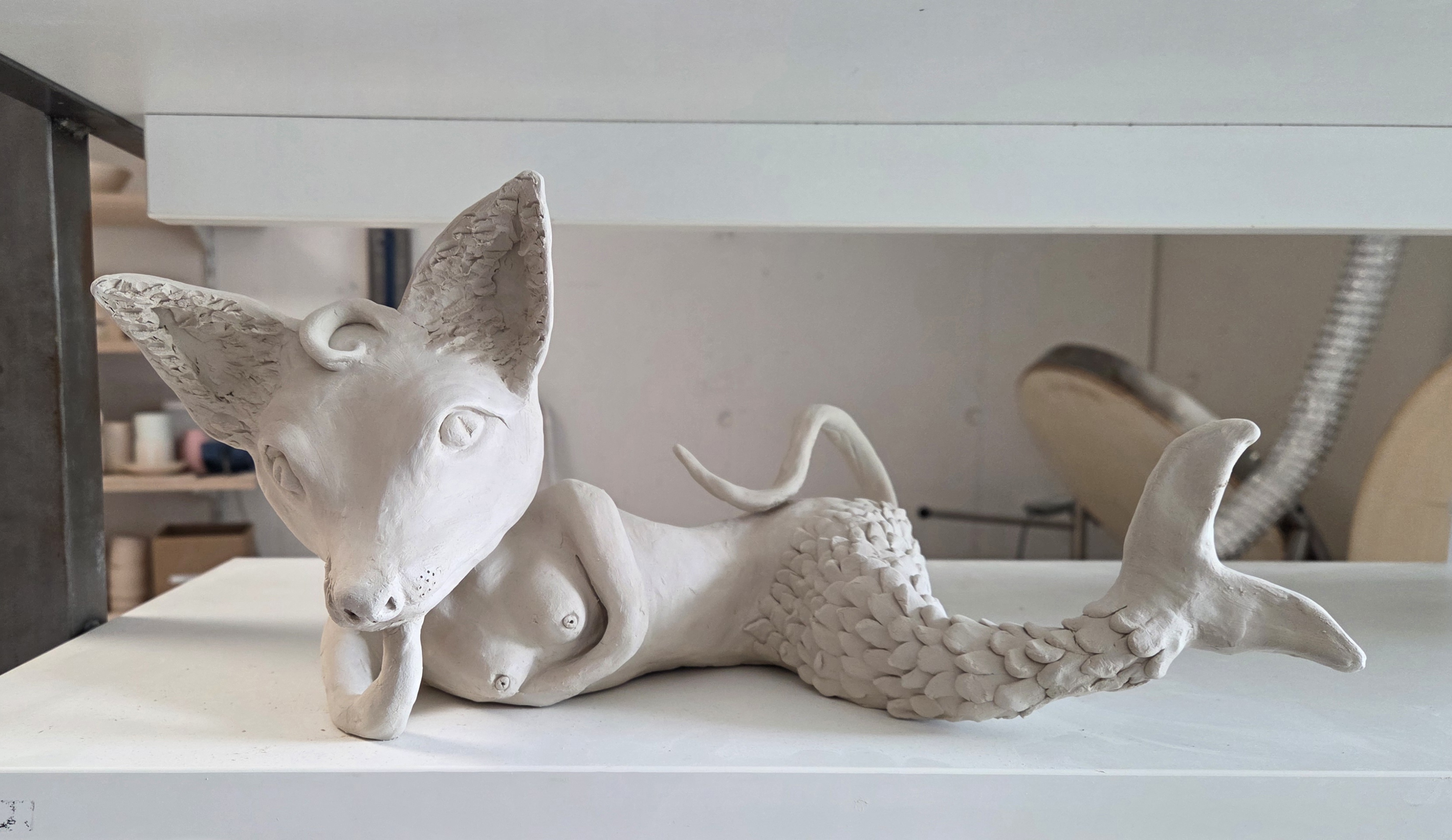 Liegende Tonskulptur einer Meerjungfrau mit Fuchskopf, aus weissem ungebranntem Ton. Grosse spitze Ohren, weiblicher Oberkörper und ein langer Fischschwanz mit detailliert modellierten Schuppen.