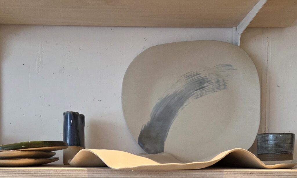Handgeformter Keramikteller mit organischer, unregelmäßiger Form, mattierter transparenter Glasur und einem abstrakten graublauen Pinselstrich, der diagonal über die Oberfläche verläuft. Der Teller lehnt in einem Regal an der Wand, umgeben von weiteren Keramikstücken.