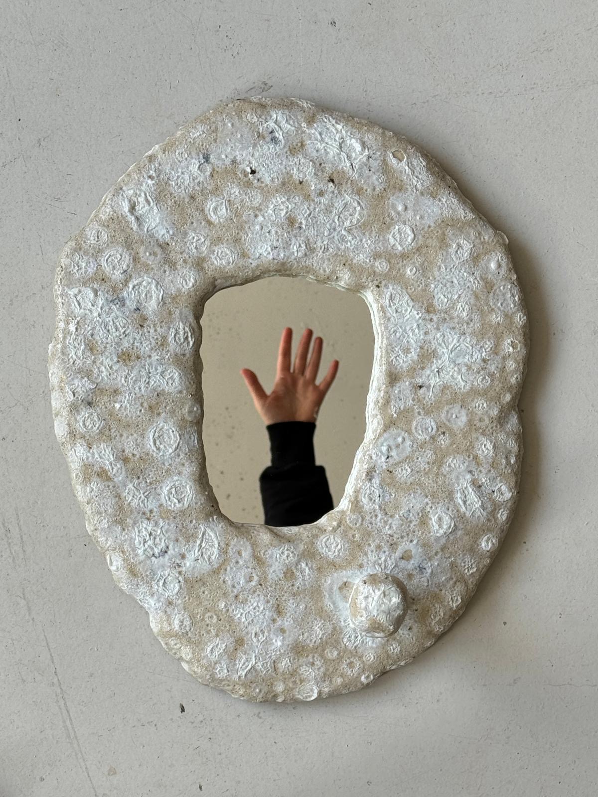 Weisser, ovaler Keramik-Wandspiegel mit strukturierter, blasiger Oberfläche und kleinem rundem Knauf, der eine ausgestreckte Hand spiegelt — Mirror, Mirror Keramik Workshop an den Zurich Design Weeks bei db pottery